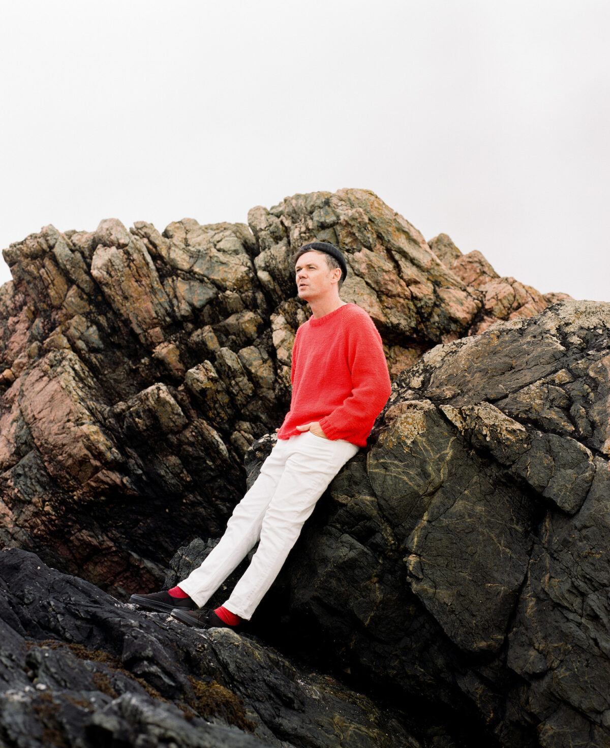 A man wearing a red jumper and white trousers leans against rocks.