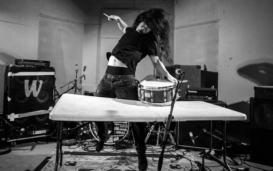 Ryosuke Kiyasu photographed jumping a short way off the ground as he prepares to hit a snare drum balanced on a folding table.