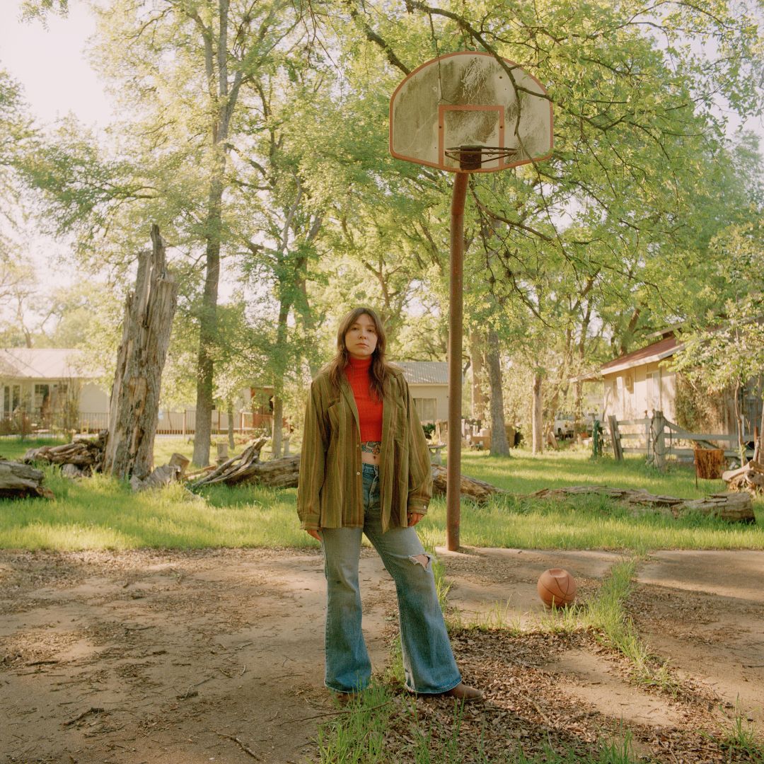 Photograph of Julianna Riolino standing on an overgrown basketball court, next to a deflated basketball and rust-covered hoop.