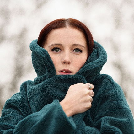 Press photo of Izzie Derry. A white woman with warm brown hair wraps herself tightly in a green coat.