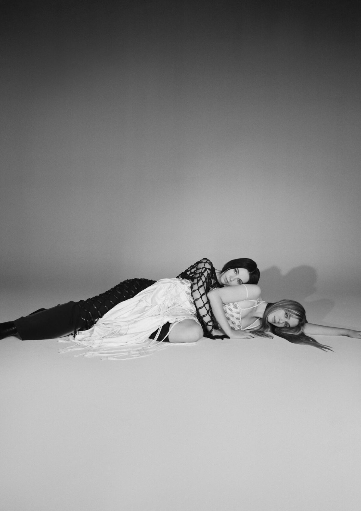 Photo of Clementine Valentine in black and white. Two women lay on the floor of a photography studio. Clementine is wearing a black dress and has black hair. She has her arms around Valentine, who is wearing a white dress.