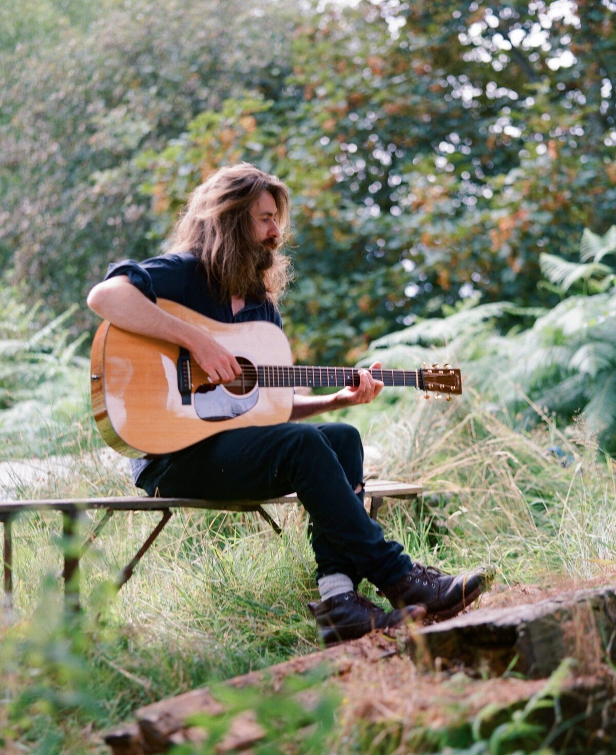 Photo of Robin Adams ahead of his show at The Tin Music and Arts on Friday 19th April 2024. Support comes from Nowhere Club and Keiron Farow. Image is of a bearded man playing an acoustic guitar whilst sitting on a bench in woodland.