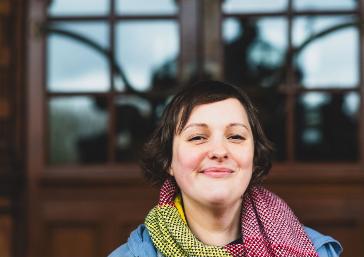 Photograph of the comedian Josie Long. A white woman with brown hair and a red and yellow scarf on top of a blue top stands in front of two large wooden doors.