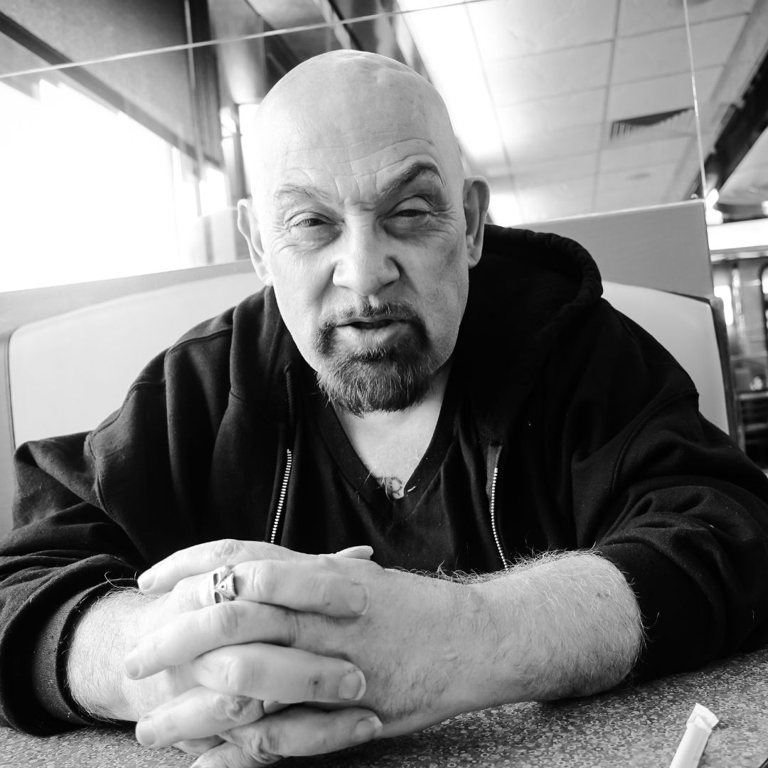 Black and white photo of Ed Hammell. A white man wearing a black hoodie is sat at a table with his hands interlaced.