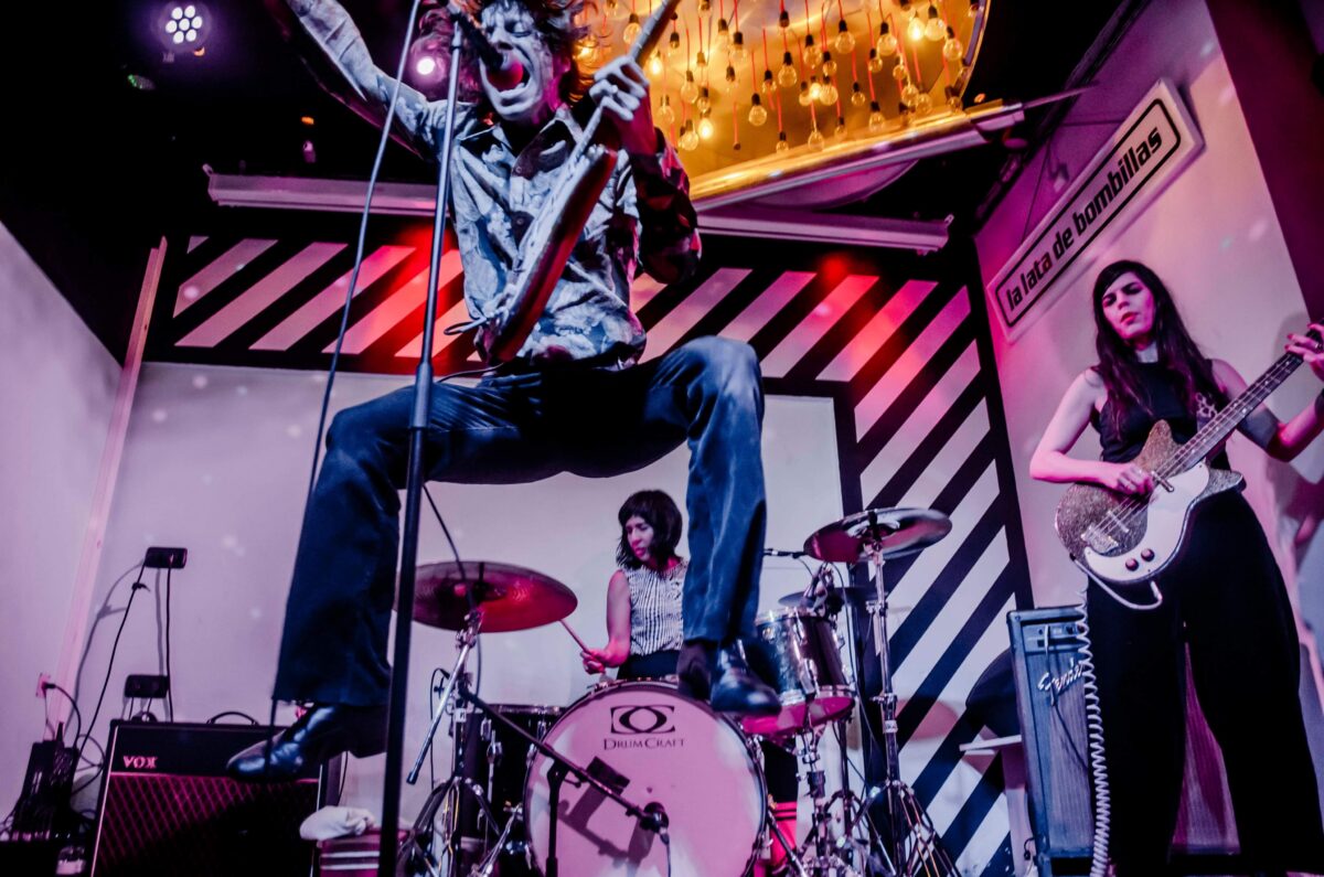 A photo of The Schizophonics live in concert. A man is singing whilst holding a guitar and jumping in the air. Behind him is a drummer. A woman stands on the right whilst playing bass guitar.