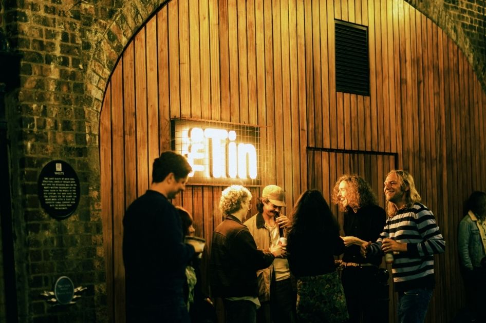 A group of people stand outside The Tin Music and Arts. A lit neon sign on the wall reads 'The Tin'.