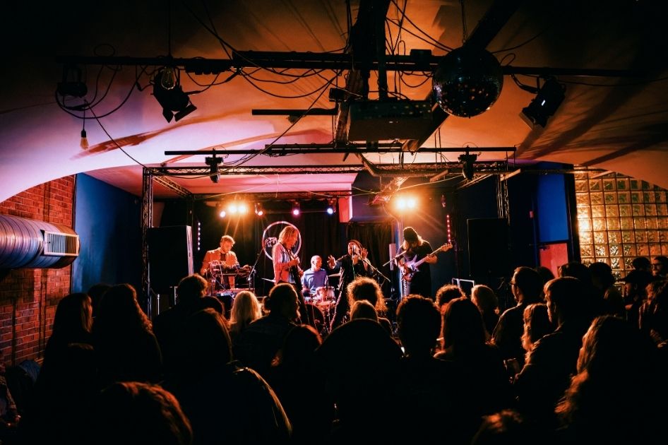 A five-piece band performs in front of an audience at The Tin Music and Arts.