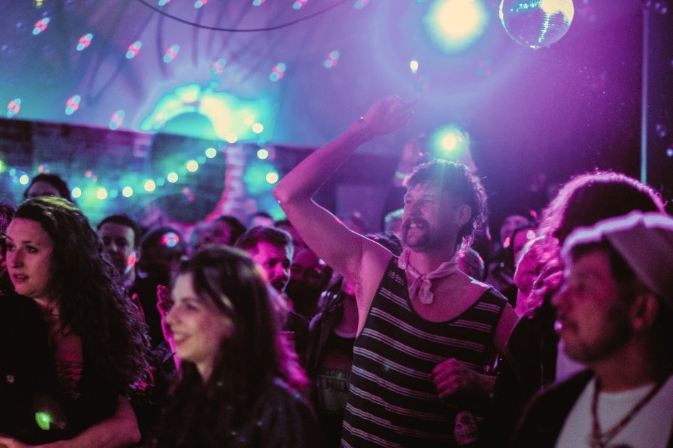 A large audience watches on at The Tin Music and Arts. There is a mirrorball hanging from the ceiling.